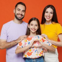 Young smiling parents mom dad with child kid daughter teen girl in basic t-shirts showing shape heart with hands heart-shape sign isolated on yellow background studio. Family day parenthood concept.