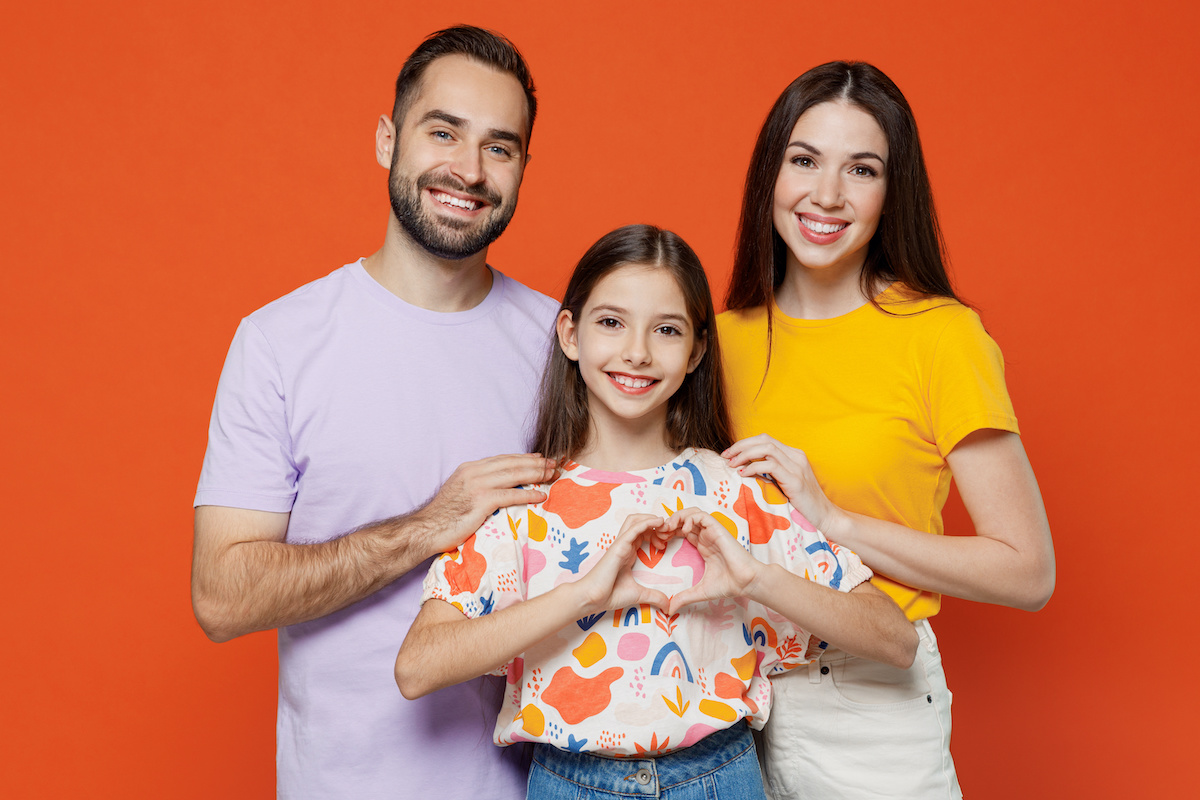 Young smiling parents mom dad with child kid daughter teen girl in basic t-shirts showing shape heart with hands heart-shape sign isolated on yellow background studio. Family day parenthood concept.