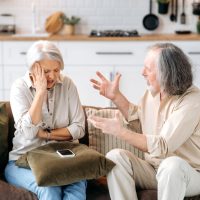 Mature caucasian couple, pensioners, sit on the sofa in the living room, quarrel with each other, shout, swear, experience irritation and indignation, disagree with each other, gesticulate with hands