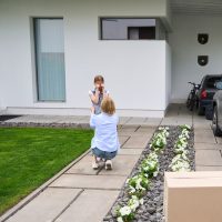 Young woman calms a crying daughter near a country house, a father carries boxes to his car Young woman calms a crying daughter near a country house, a father carries boxes to his car