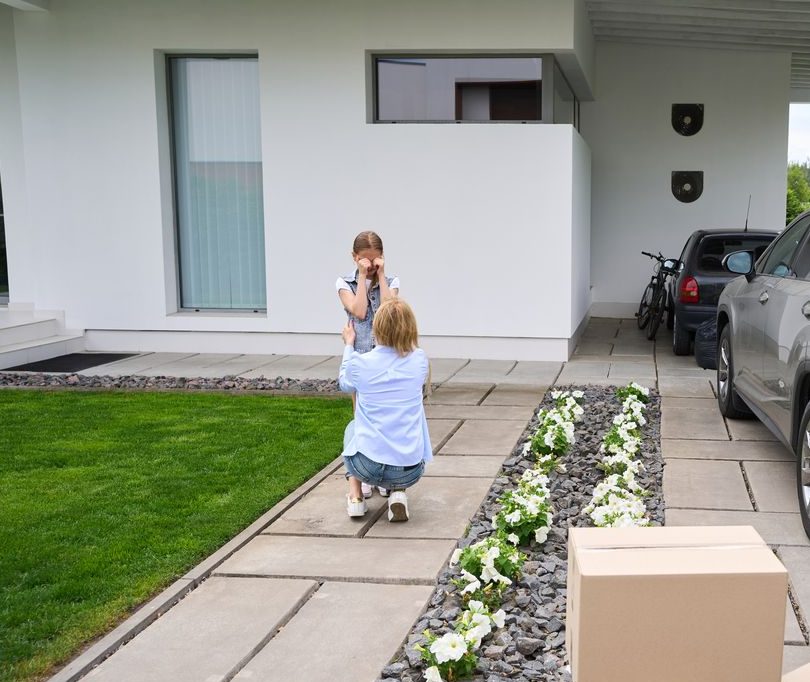 Young woman calms a crying daughter near a country house, a father carries boxes to his car