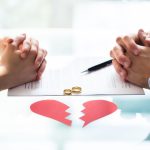 Couple's Hand On Divorce Paper With Wedding Rings And Broken Heart