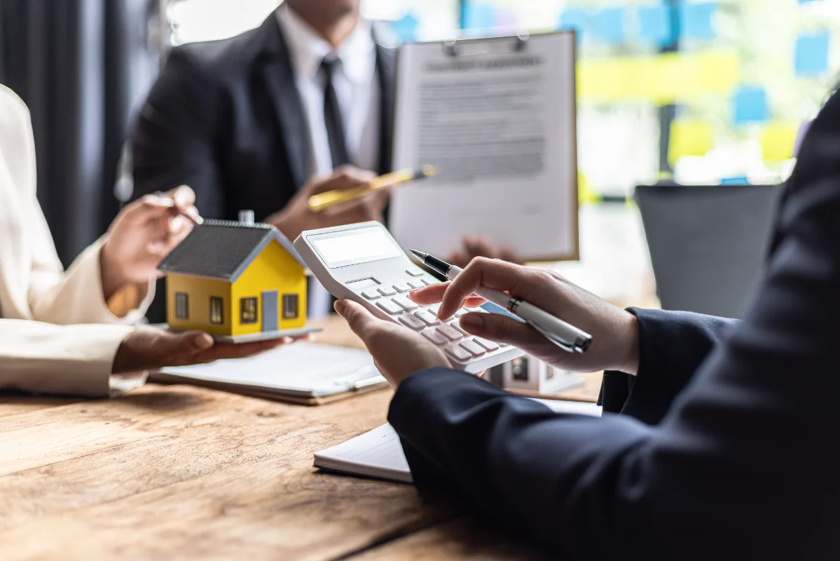 Professionals meet at a table to review documents, with one person using a calculator and a small model house nearby.