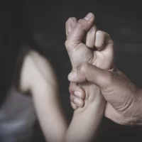 A hand grips another person's wrist tightly against a dark background, conveying a tense or controlling situation. The mood is somber and intense.