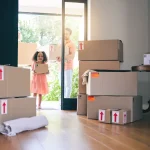 a-little-girl-and-an-adult-carry-boxes-into-a-bright-living-room-filled-with-moving-boxes